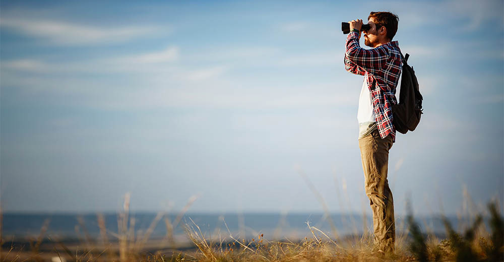 Een man die tijdens een wandeling in de duinen met verrekijker uitkijkend over de natuur. - Oxtarn Clinics & Adventures