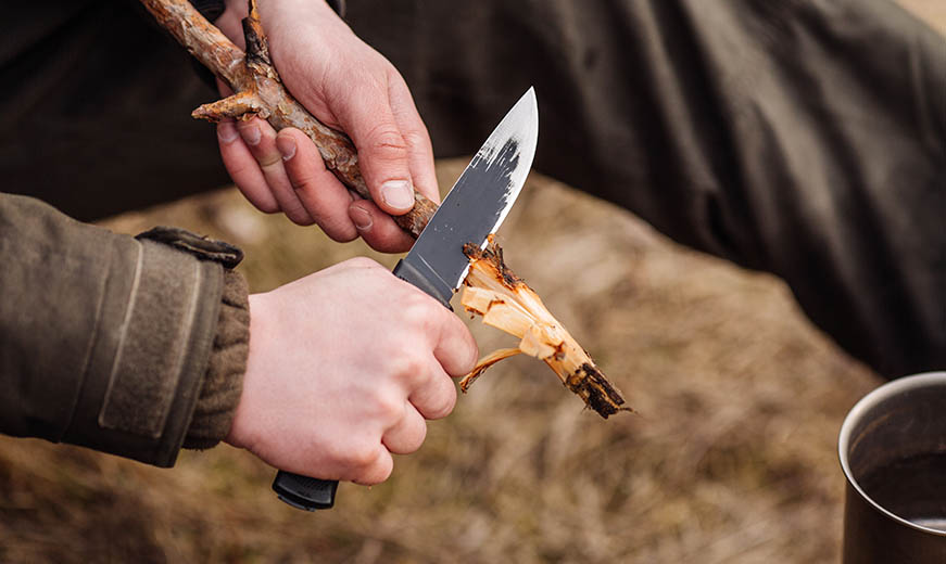 Man met mes bezig met het maken van geveerde stokken voor een kampvuur tijdens een survival clinic. - Oxtarn Clinics & Adventures