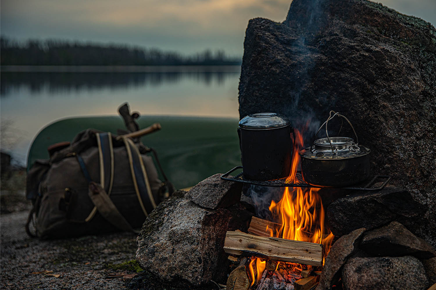 Kampeer survival avontuur bij het meer met pannen boven een kampvuur en een tas tegen de rotsen. - Oxtarn Clinics & Adventures